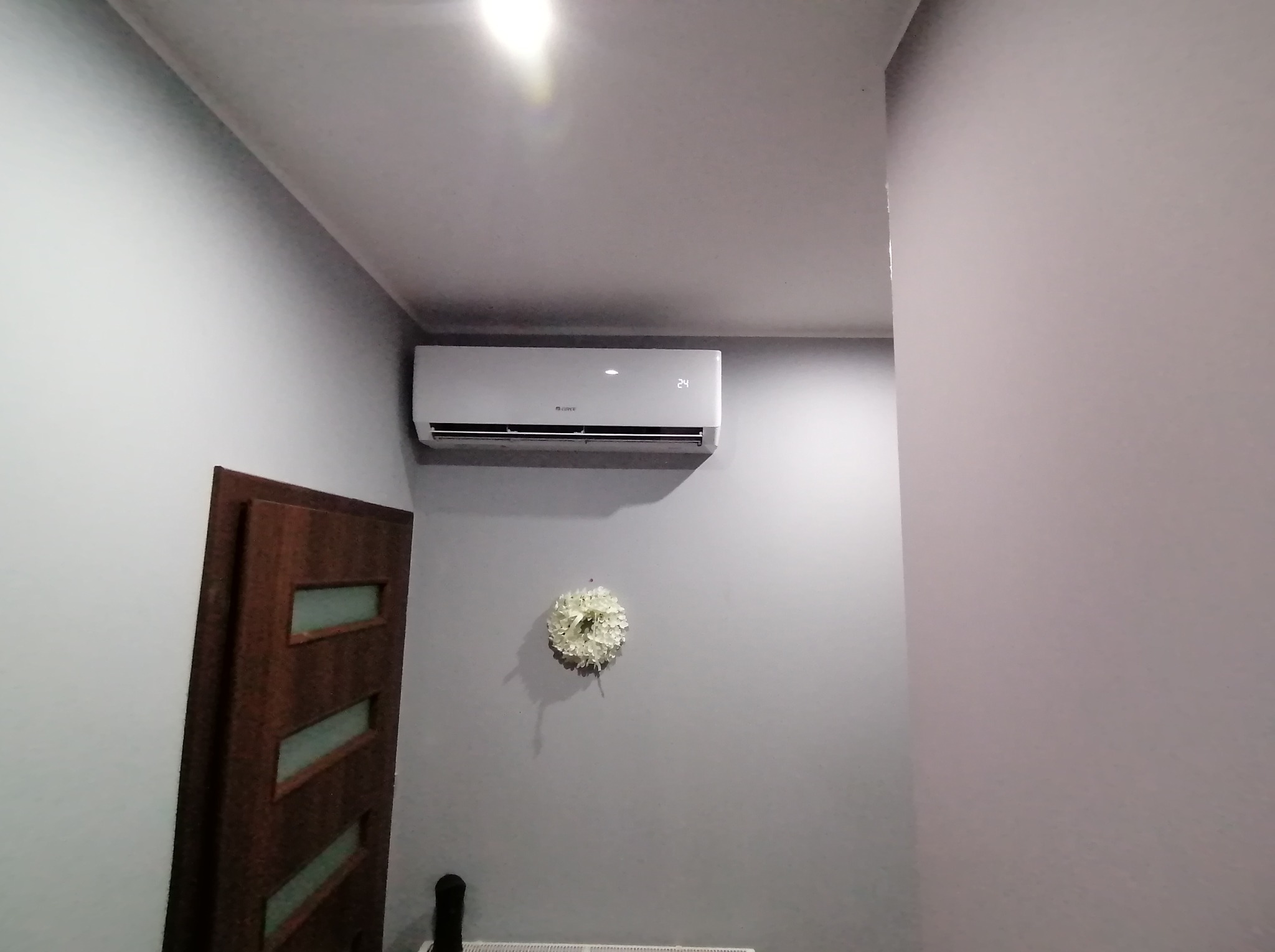 Biała jednostka klimatyzacyjna zamontowana na szarej ścianie w pokoju, widoczny wyświetlacz z temperaturą 24 stopnie, obok drewniane drzwi z szybami i wieniec dekoracyjny.