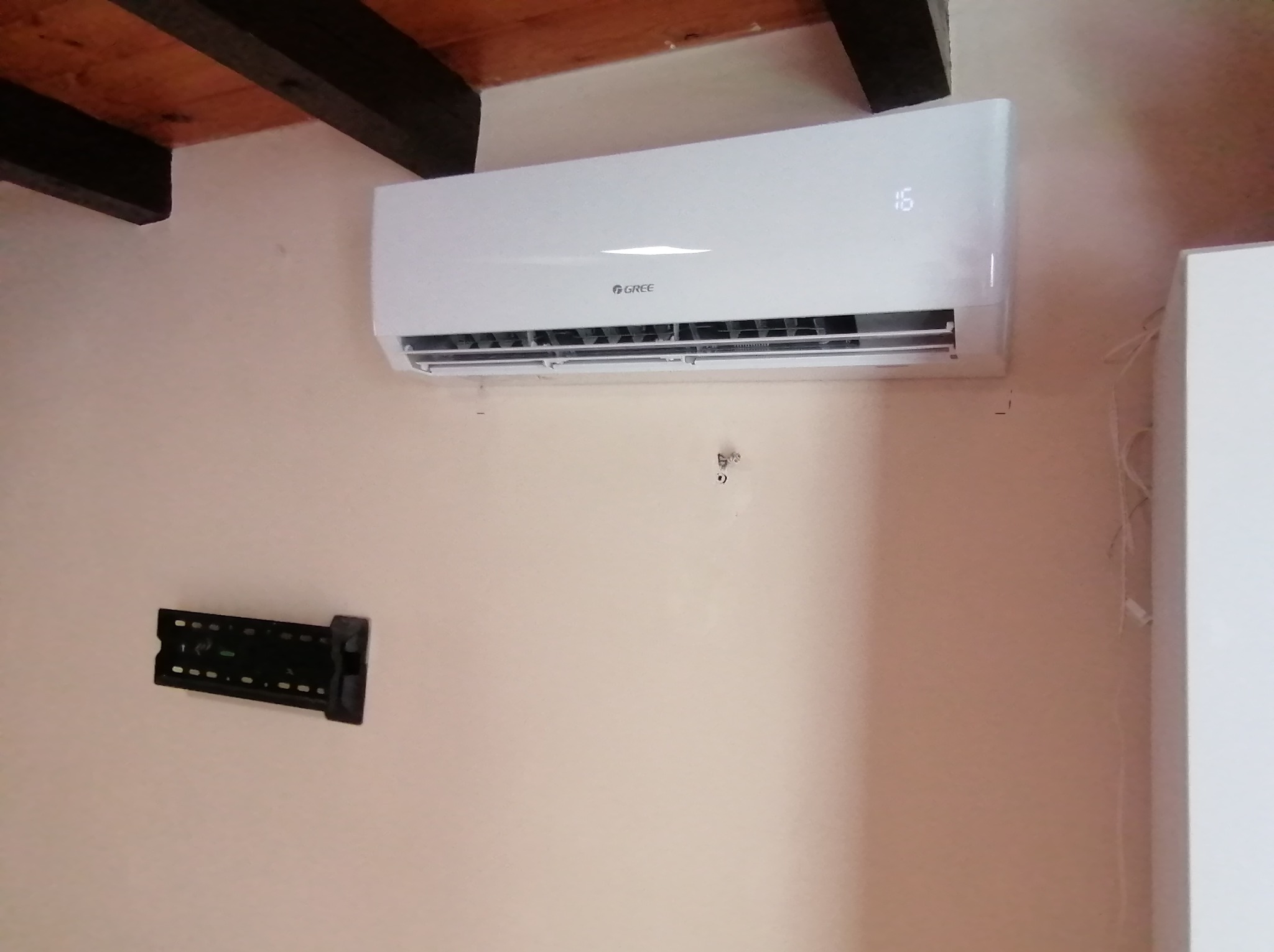 Biały klimatyzator marki Gree zamontowany na ścianie w pomieszczeniu z drewnianym sufitem, obok widoczny uchwyt do telewizora.