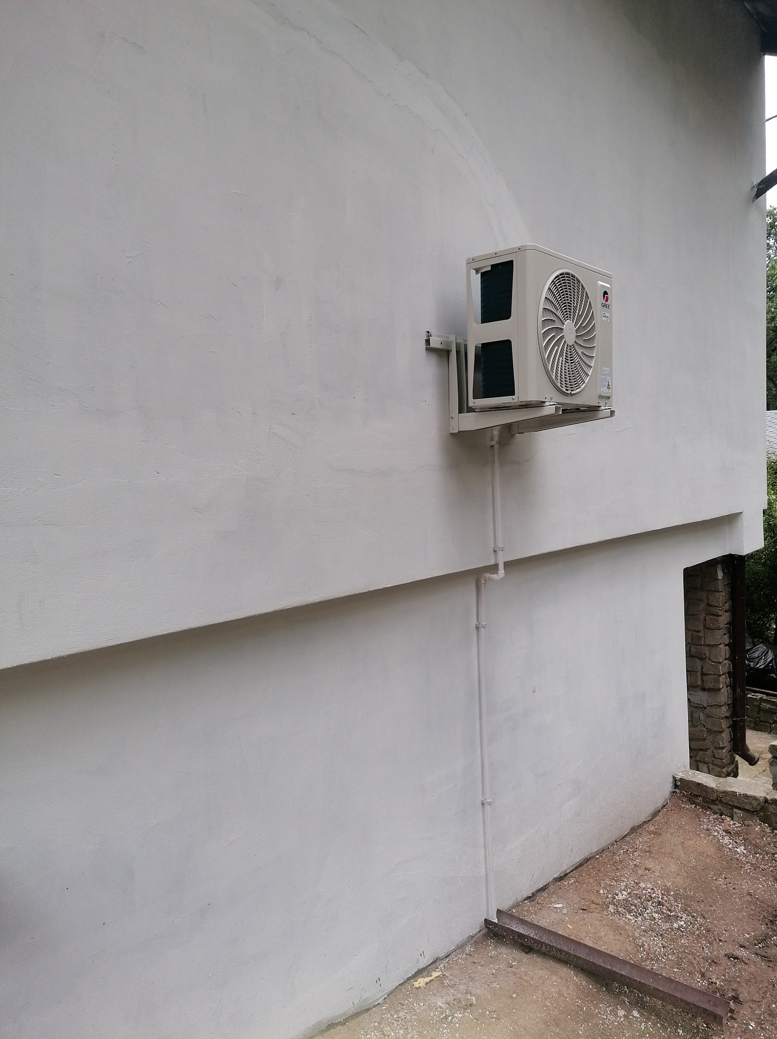Zewnętrzna jednostka klimatyzacji marki Gree zamontowana na wsporniku na szarej, chropowatej ścianie budynku, z widocznymi rurami odprowadzającymi skropliny.