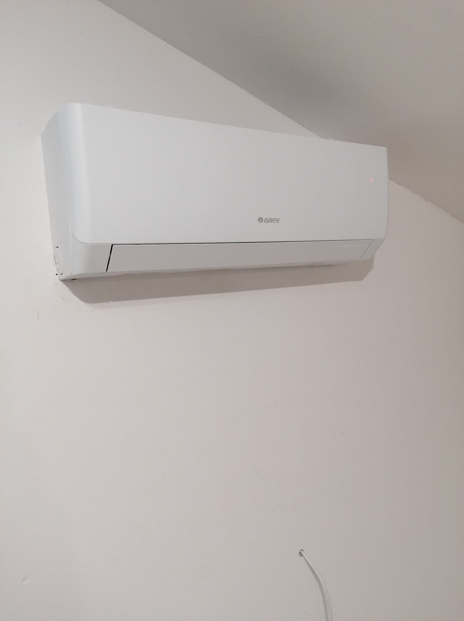 Biały klimatyzator marki Gree zamontowany na jasnej ścianie, widoczny delikatny kabel wystający spod urządzenia.