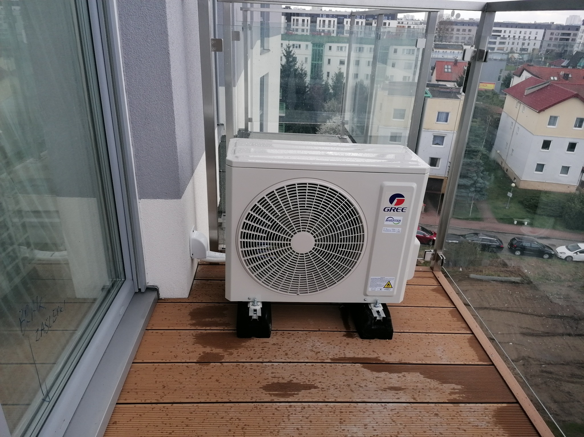Jednostka zewnętrzna klimatyzatora GREE zamontowana na balkonie z drewnianą podłogą kompozytową, widok na osiedle mieszkaniowe przez szklaną balustradę.
