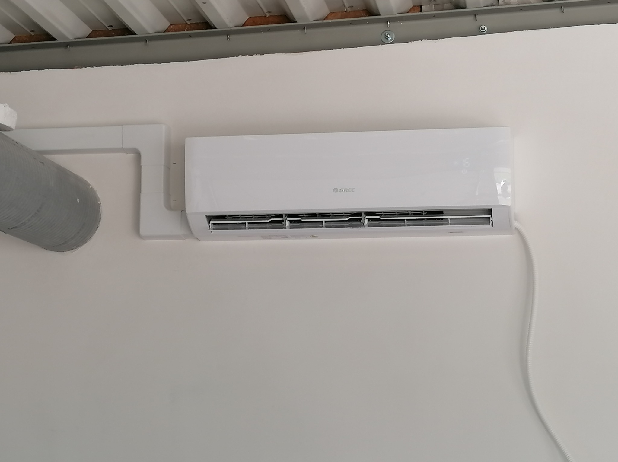Biała jednostka wewnętrzna klimatyzatora marki Gree zamontowana na białej ścianie pod metalową konstrukcją sufitu, z widoczną szarą rurą wentylacyjną po lewej stronie i kablem zasilającym po prawej.