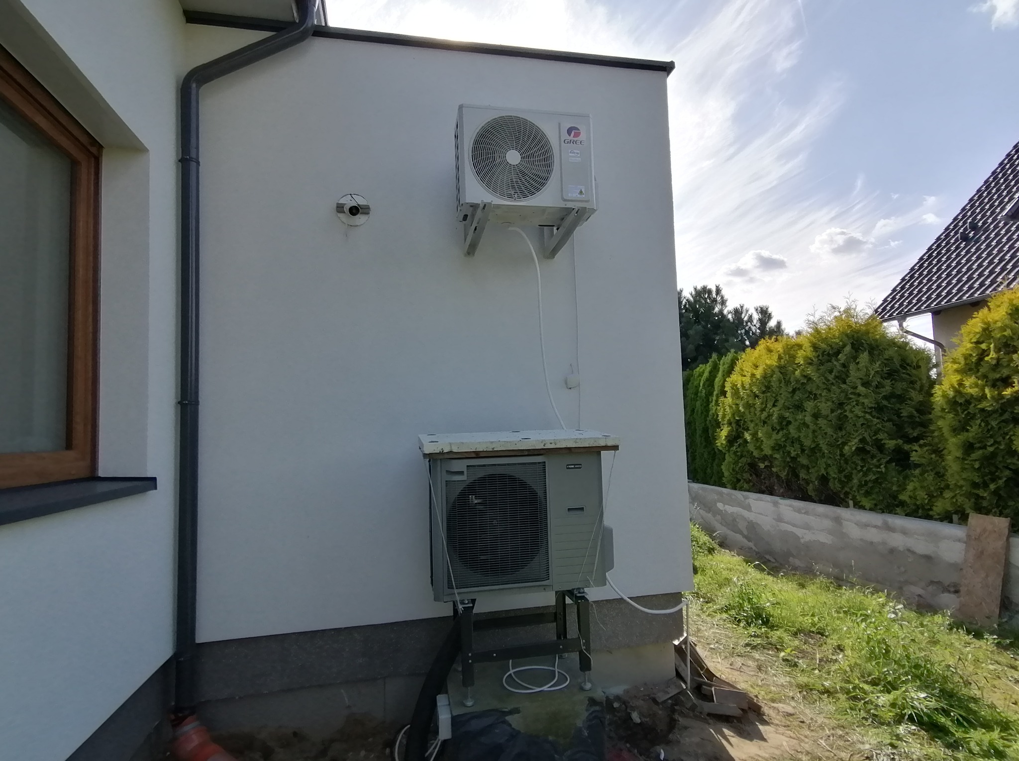 Zewnętrzna jednostka klimatyzacji marki Gree zamontowana na białej ścianie domu, z widocznymi przewodami łączącymi ją z jednostką stojącą na ziemi na metalowej konstrukcji, na tle błękitnego nieba...
