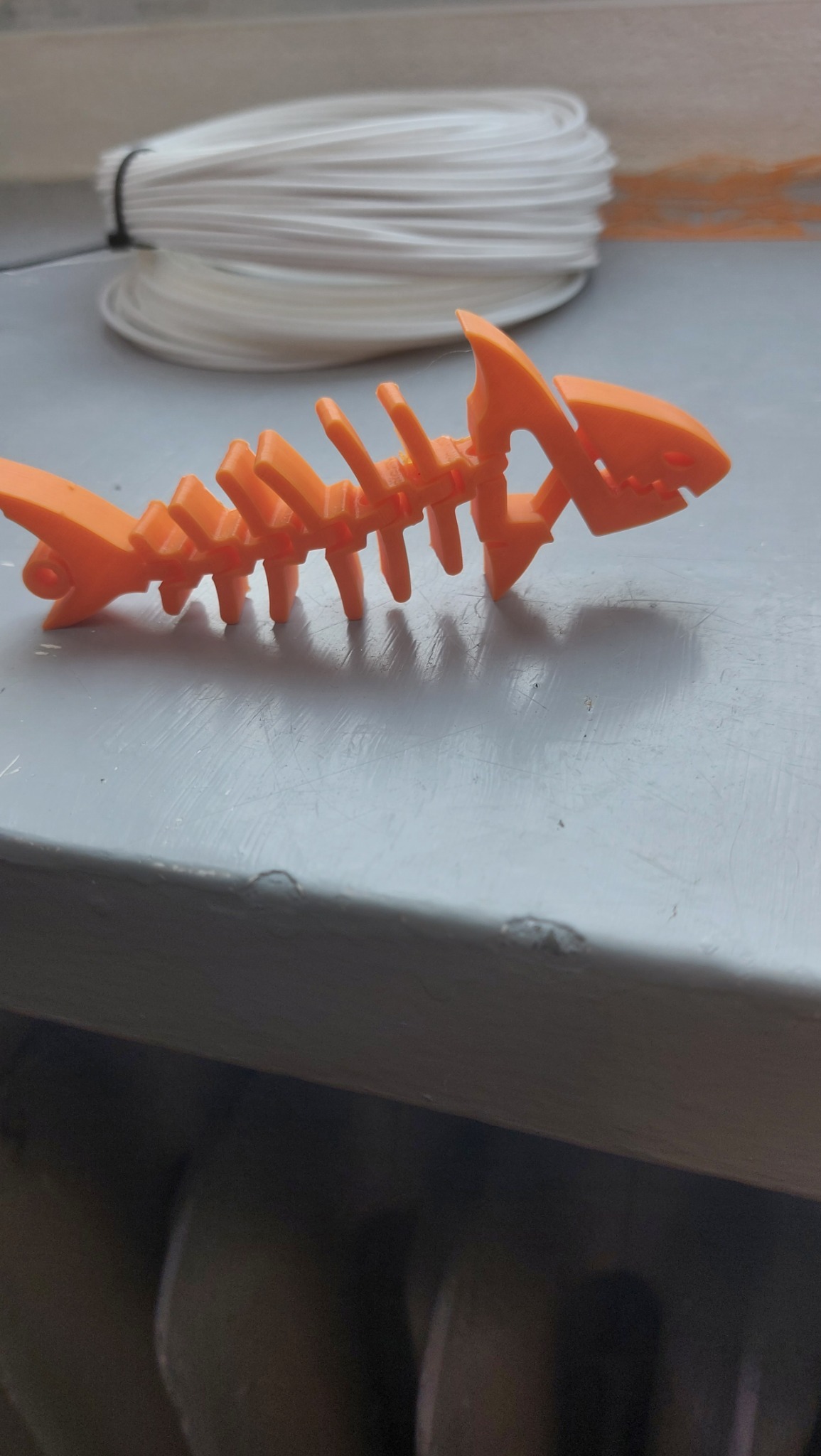 Pomarańczowy szkielet ryby wydrukowany w 3D, leżący na szarej powierzchni, z rolką białego filamentu w tle.