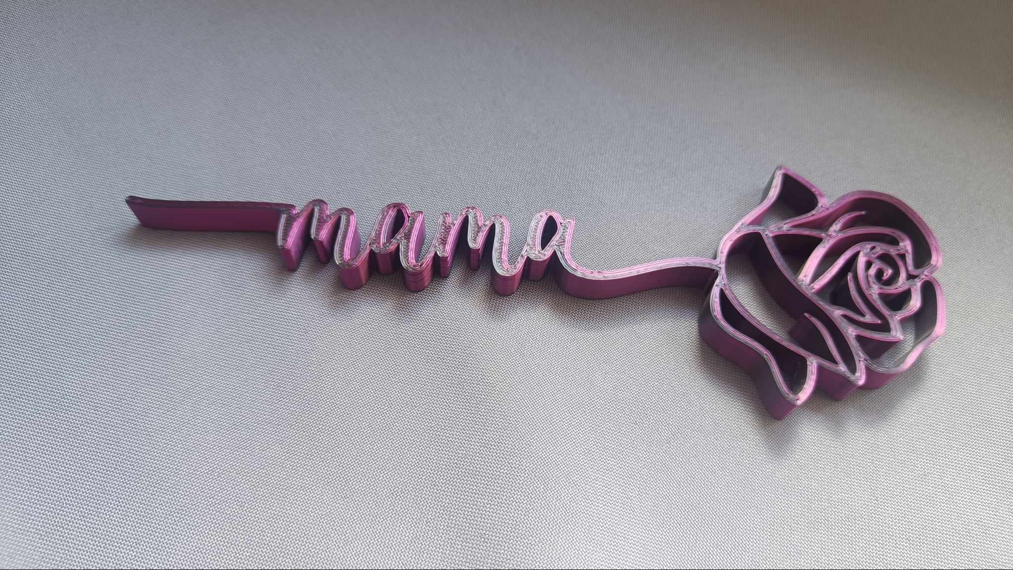 Różowa, przestrzenna forma wycięta laserowo, przedstawiająca napis 'mama' połączony z motywem róży, leżąca na szarym tle.