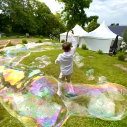 Chłopiec w białej koszuli puszcza ogromne bańki mydlane na trawie, w tle białe namioty i drzewa.