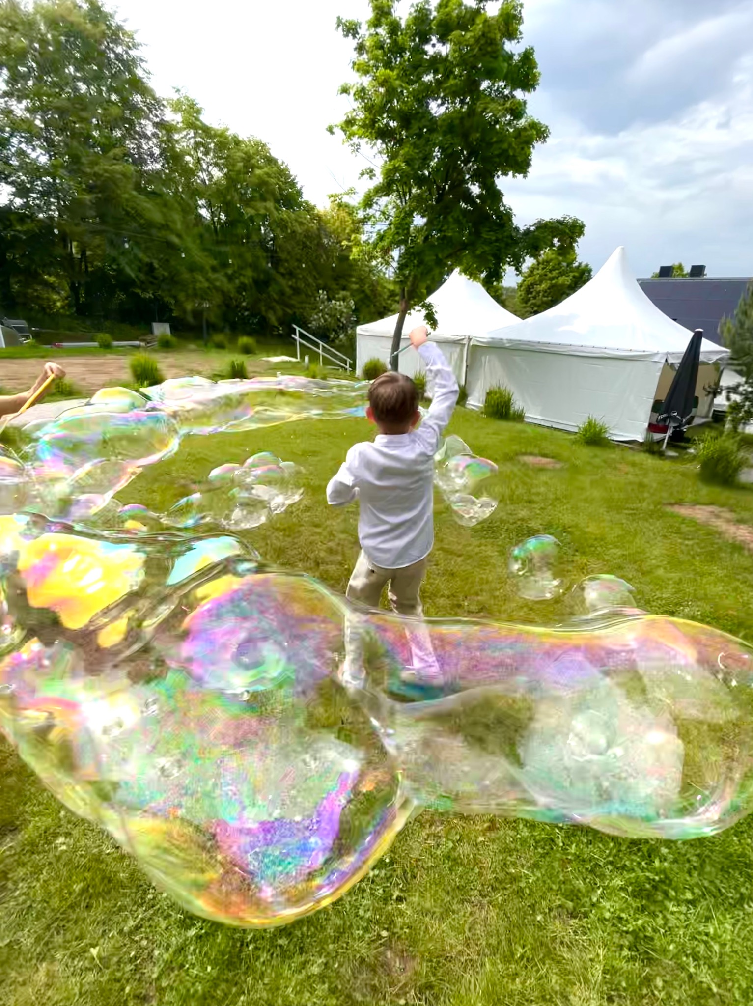Chłopiec w białej koszuli puszcza ogromne bańki mydlane na trawie, w tle białe namioty i drzewa.