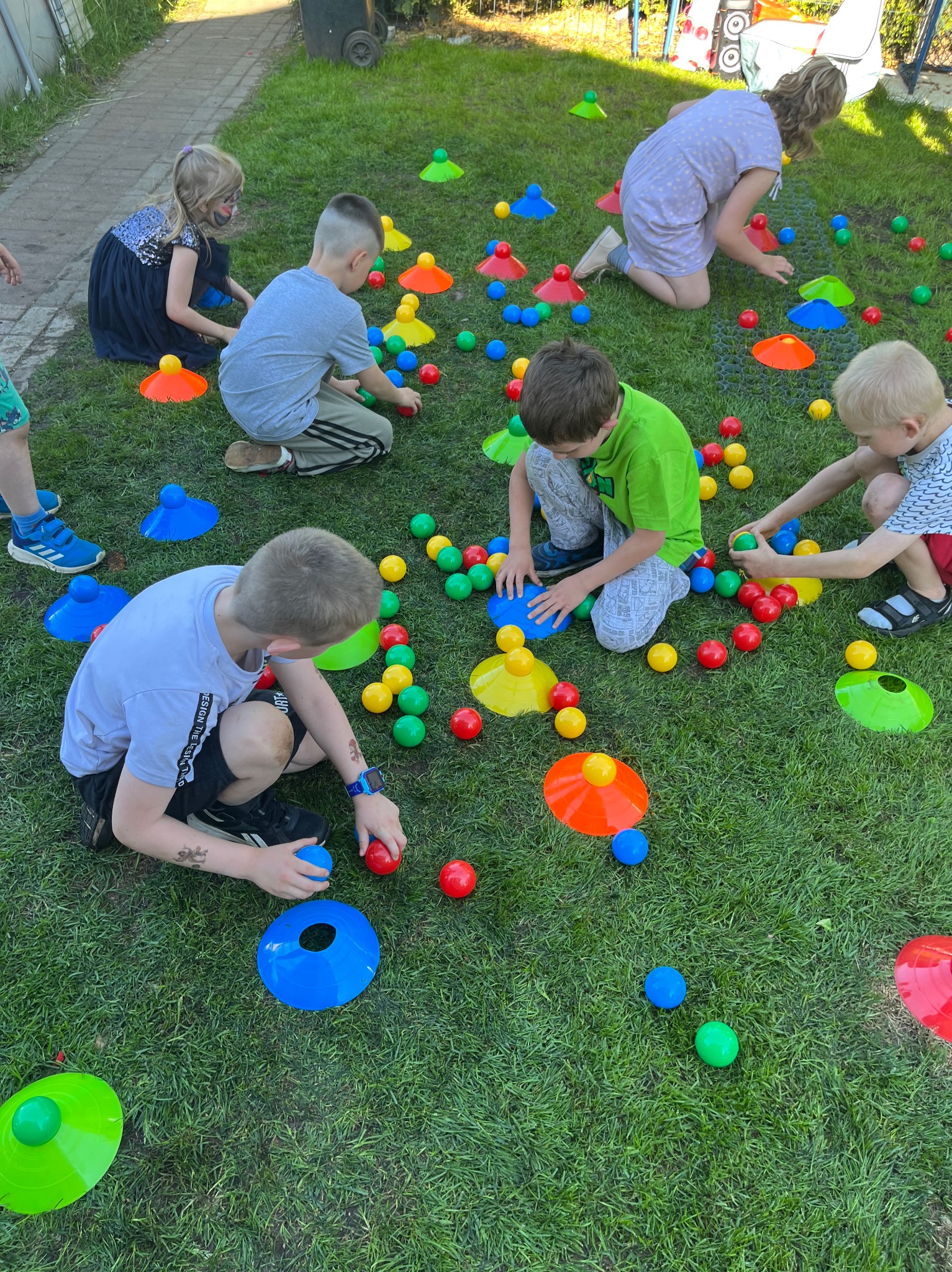 Grupa dzieci siedzi na trawie podczas zabawy z kolorowymi piłkami i pachołkami, tworząc radosną atmosferę na świeżym powietrzu.