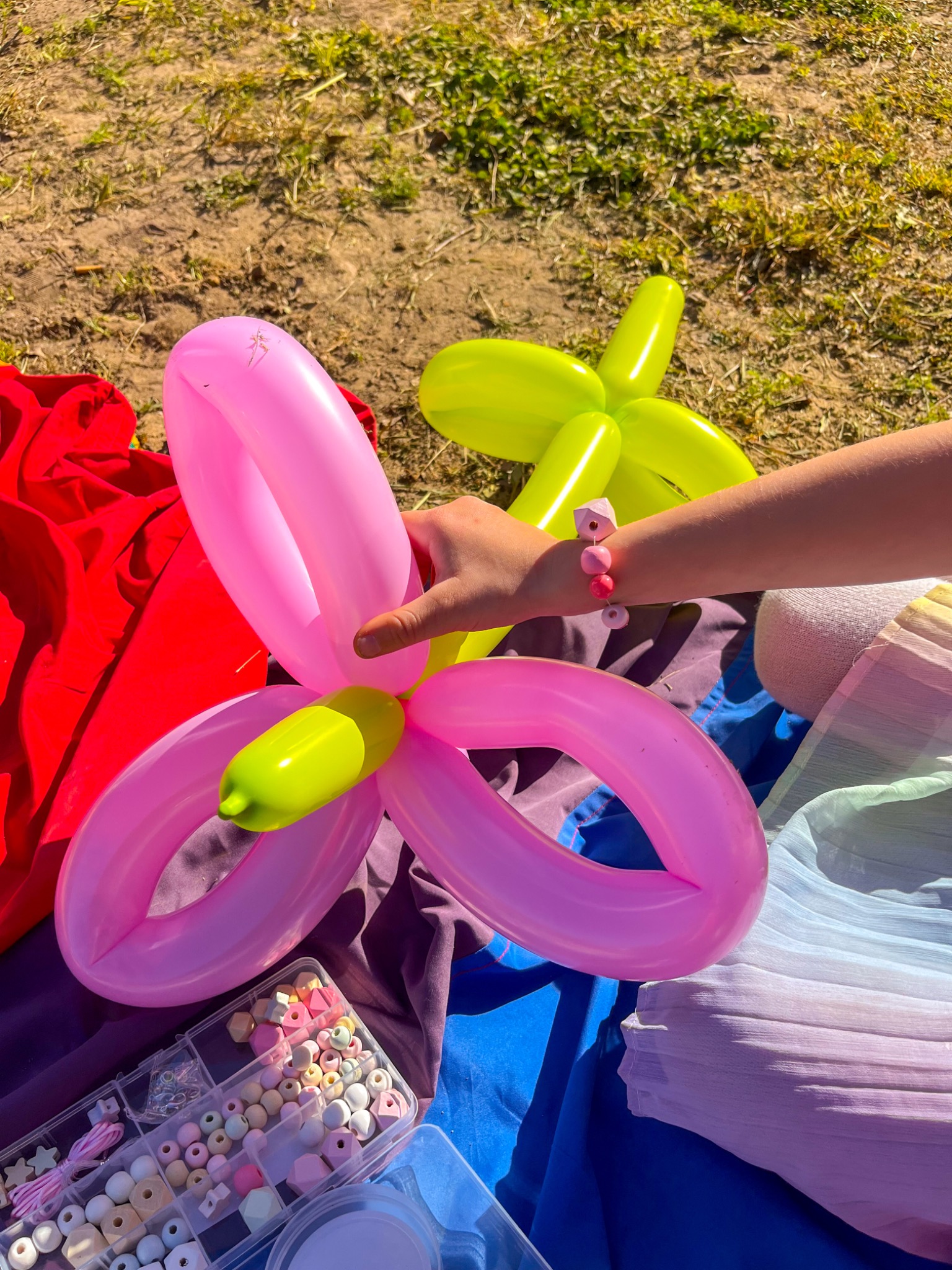 Ręka dziecka formuje różowego balonowego kwiatka z zielonym środkiem, obok leży drugi, zielony kwiatek z balonów, pudełko z koralikami i kolorowe tkaniny.