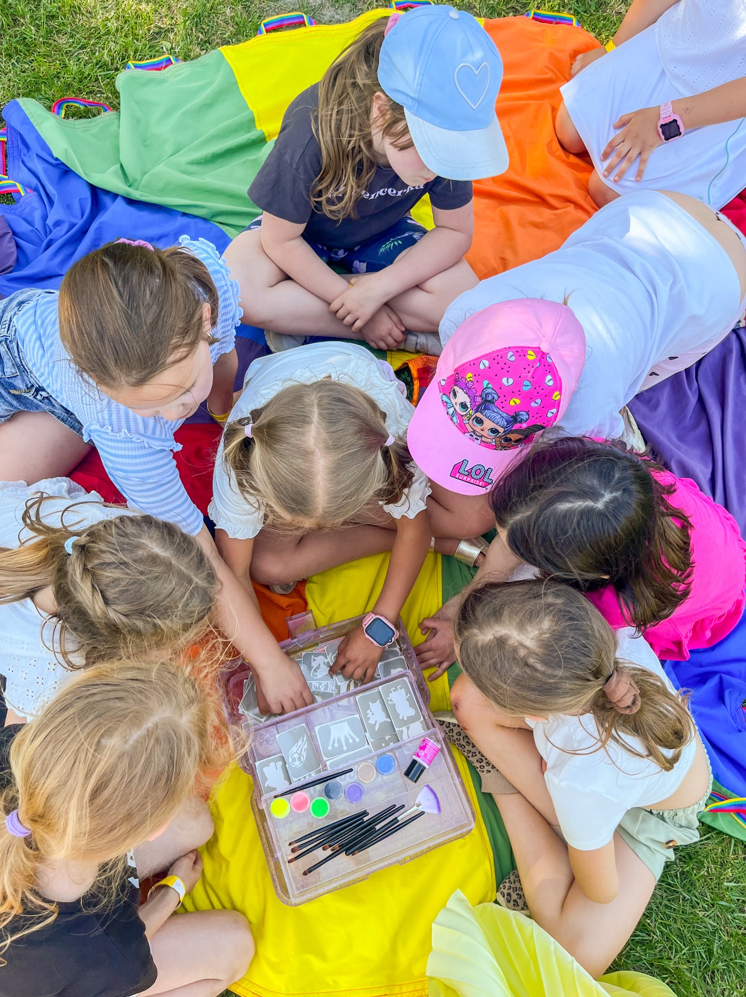 Grupa dziewczynek siedzi na kolorowej chuście na trawie, pochylona nad przezroczystym pudełkiem z farbkami i pędzelkami do malowania twarzy, przygotowując się do zabawy.