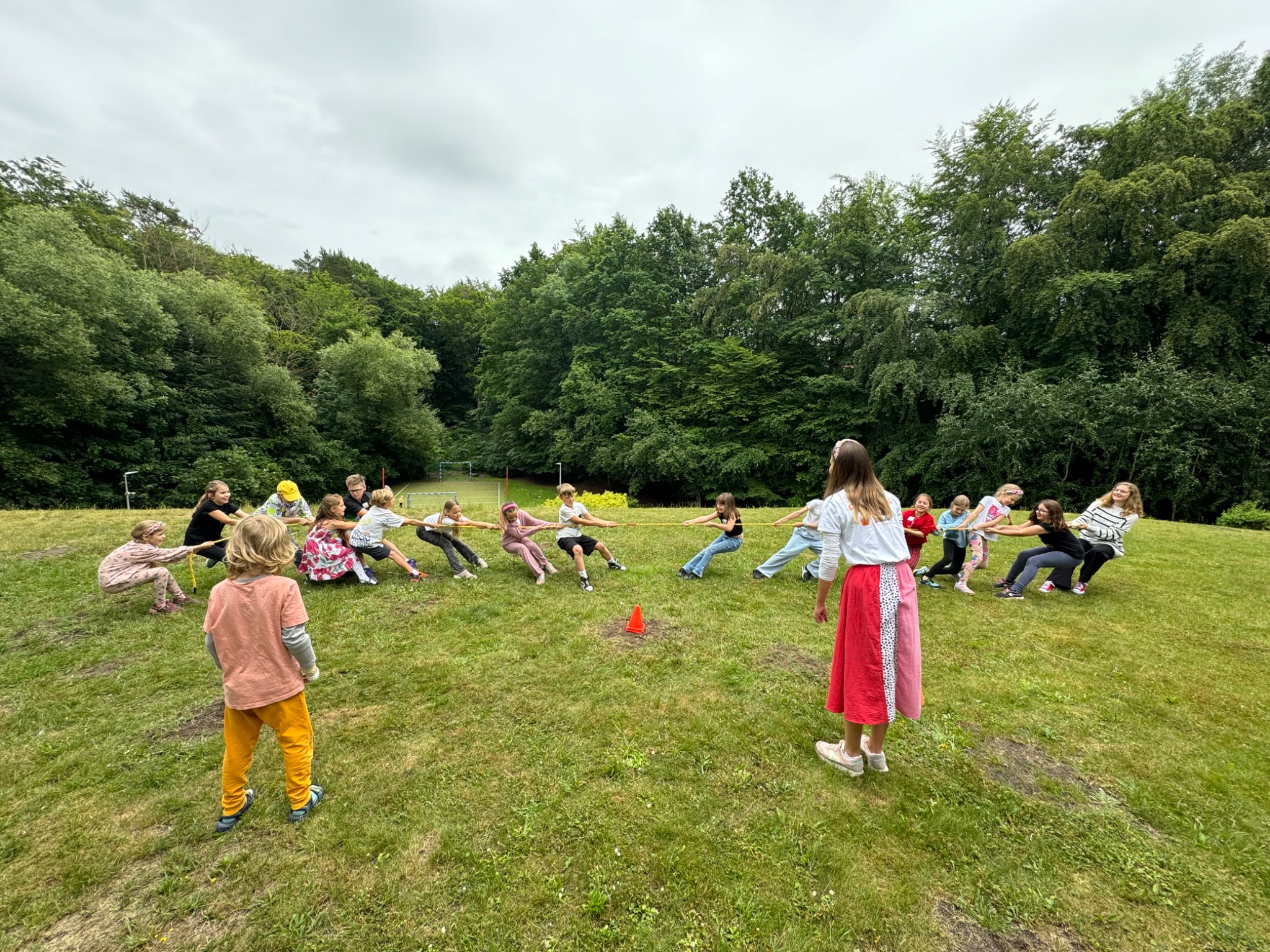 Grupa dzieci gra w przeciąganie liny na trawiastym terenie, z lasem w tle, nadzorowana przez osobę dorosłą w czerwonej spódnicy.