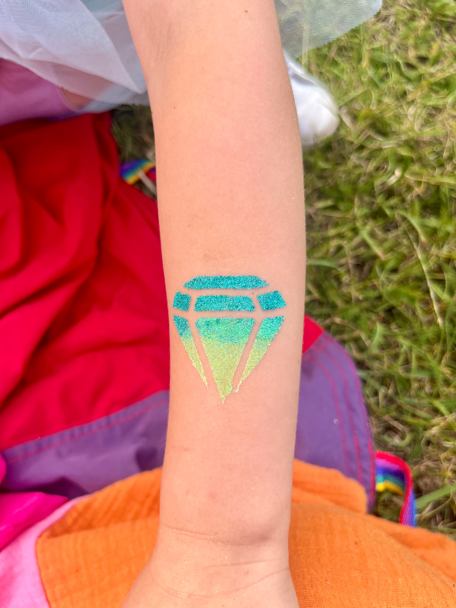 Dziecięca ręka z brokatowym tatuażem w kształcie diamentu, z gradientem kolorów od turkusowego do jasnozielonego, na tle trawy i kolorowych ubrań.