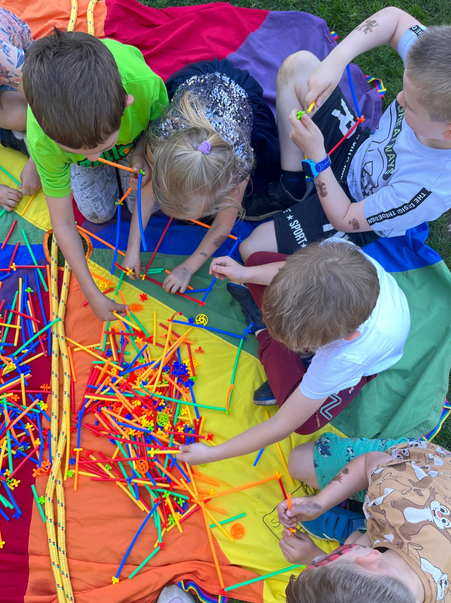 Grupa dzieci siedzi na kolorowej chuście animacyjnej, bawiąc się kolorowymi patyczkami i łącznikami, tworząc konstrukcje.