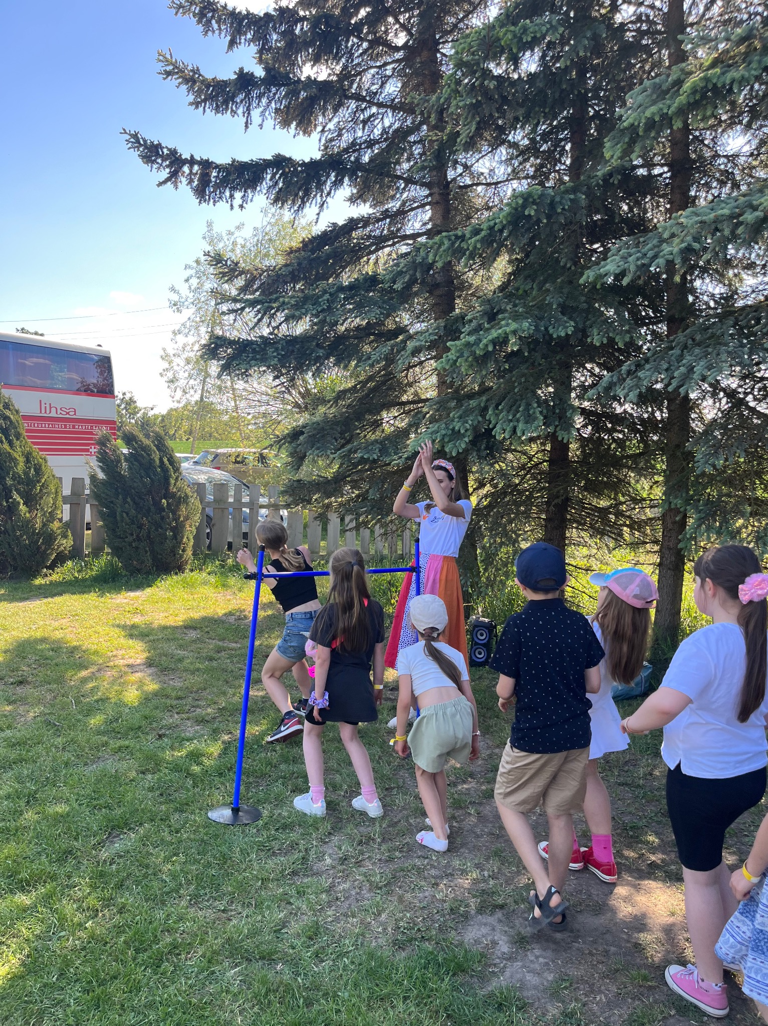 Dzieci biorące udział w zabawie z animatorem na świeżym powietrzu, widoczny autobus w tle.