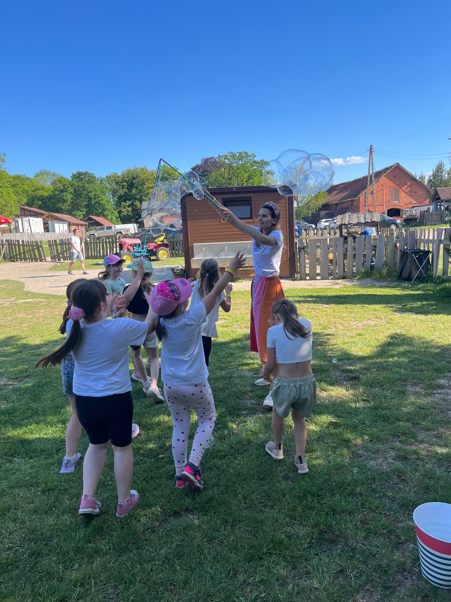 Animator tworzy duże bańki mydlane dla grupy dzieci na trawniku w słoneczny dzień, w tle zabudowania gospodarcze.