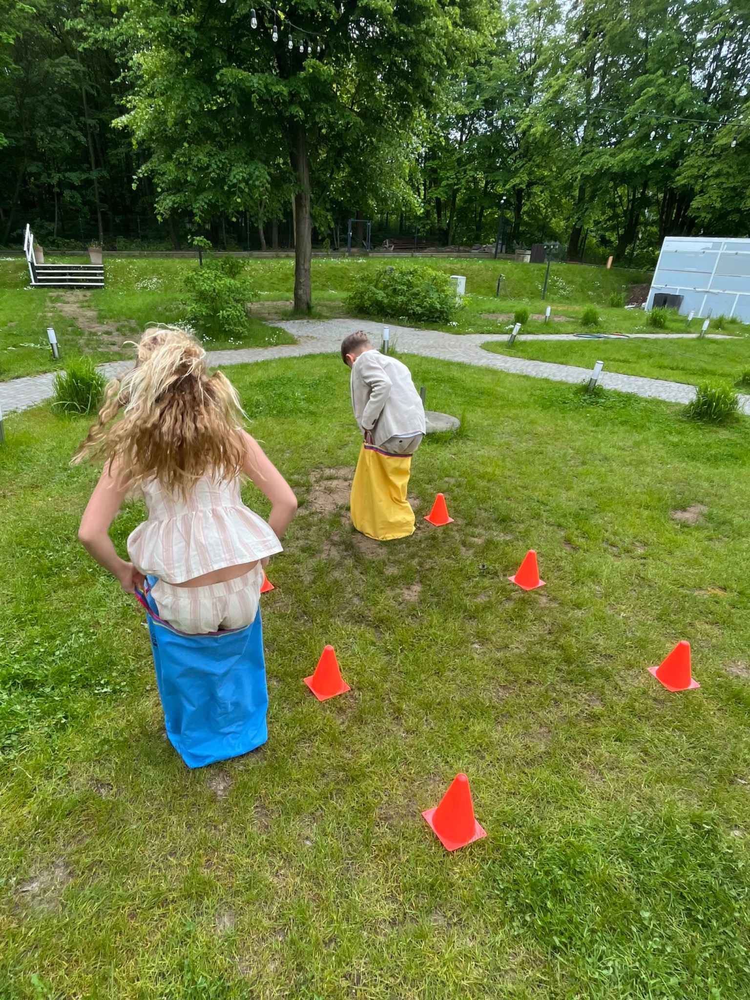 Dzieci w workach skaczą przez trawnik wyznaczony pomarańczowymi pachołkami podczas zabawy na świeżym powietrzu, w tle zieleń drzew.