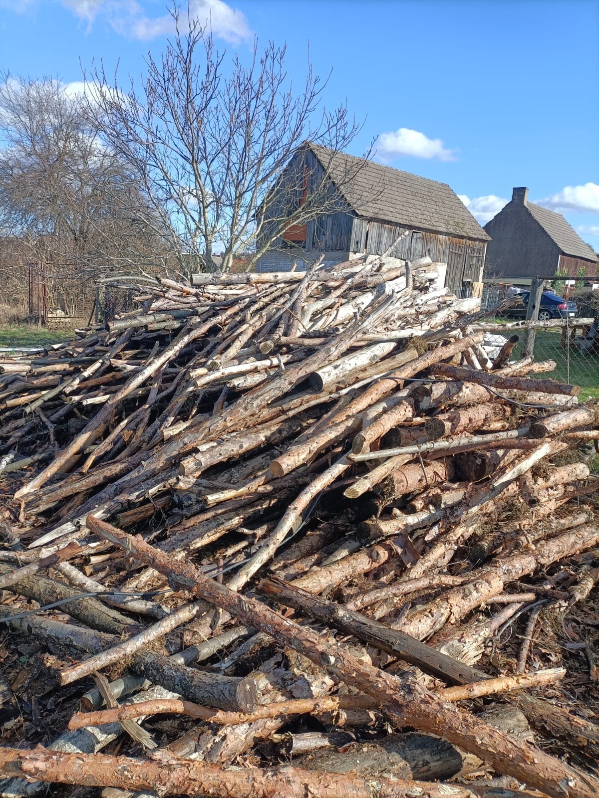 Stos pociętych gałęzi i kłód na tle wiejskiego krajobrazu z drewnianymi zabudowaniami i błękitnym niebem.
