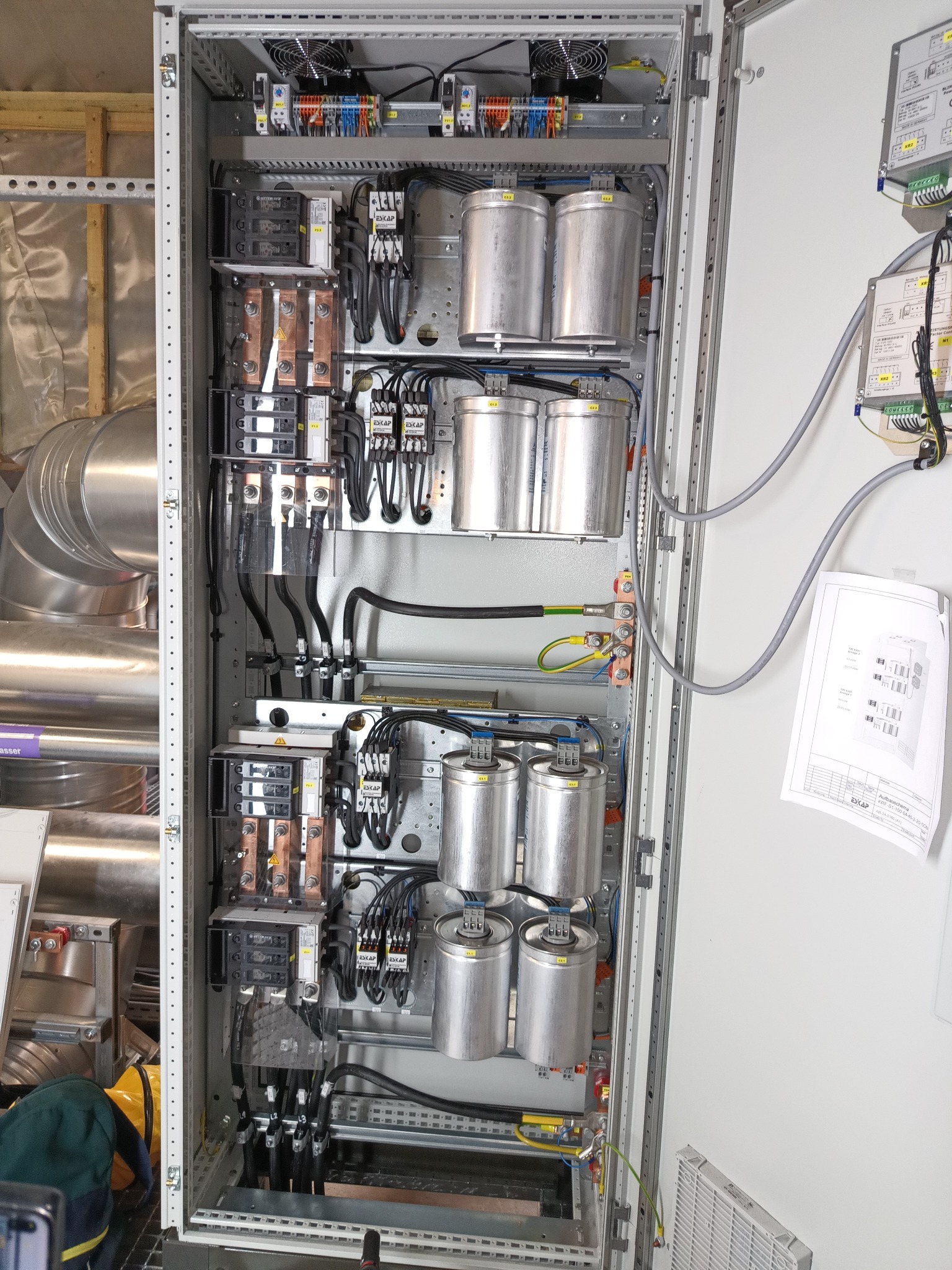 Otwarta szafa sterownicza z widocznymi kondensatorami cylindrycznymi, bezpiecznikami, okablowaniem, szynami zbiorczymi i wentylatorami chłodzącymi; schemat podłączenia na białej ścianie obok.