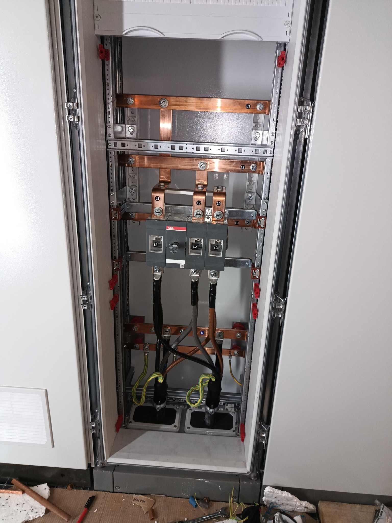 Otwarta szafa elektryczna z zamontowanymi wyłącznikami ABB, miedzianymi szynami zbiorczymi i przewodami w izolacji, widoczne oznaczenia L1, L2, L3, N, na podłodze resztki materiałów instalacyjnych...