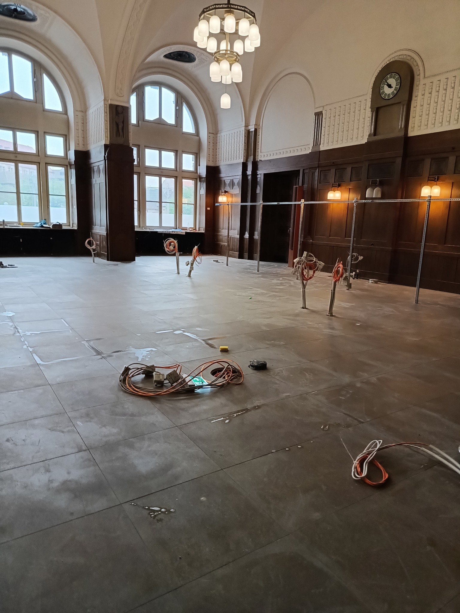 Wnętrze remontowanego pomieszczenia z wysokimi oknami i drewnianą boazerią, widoczne przewody elektryczne wystające z podłogi i zwój kabli na posadzce.