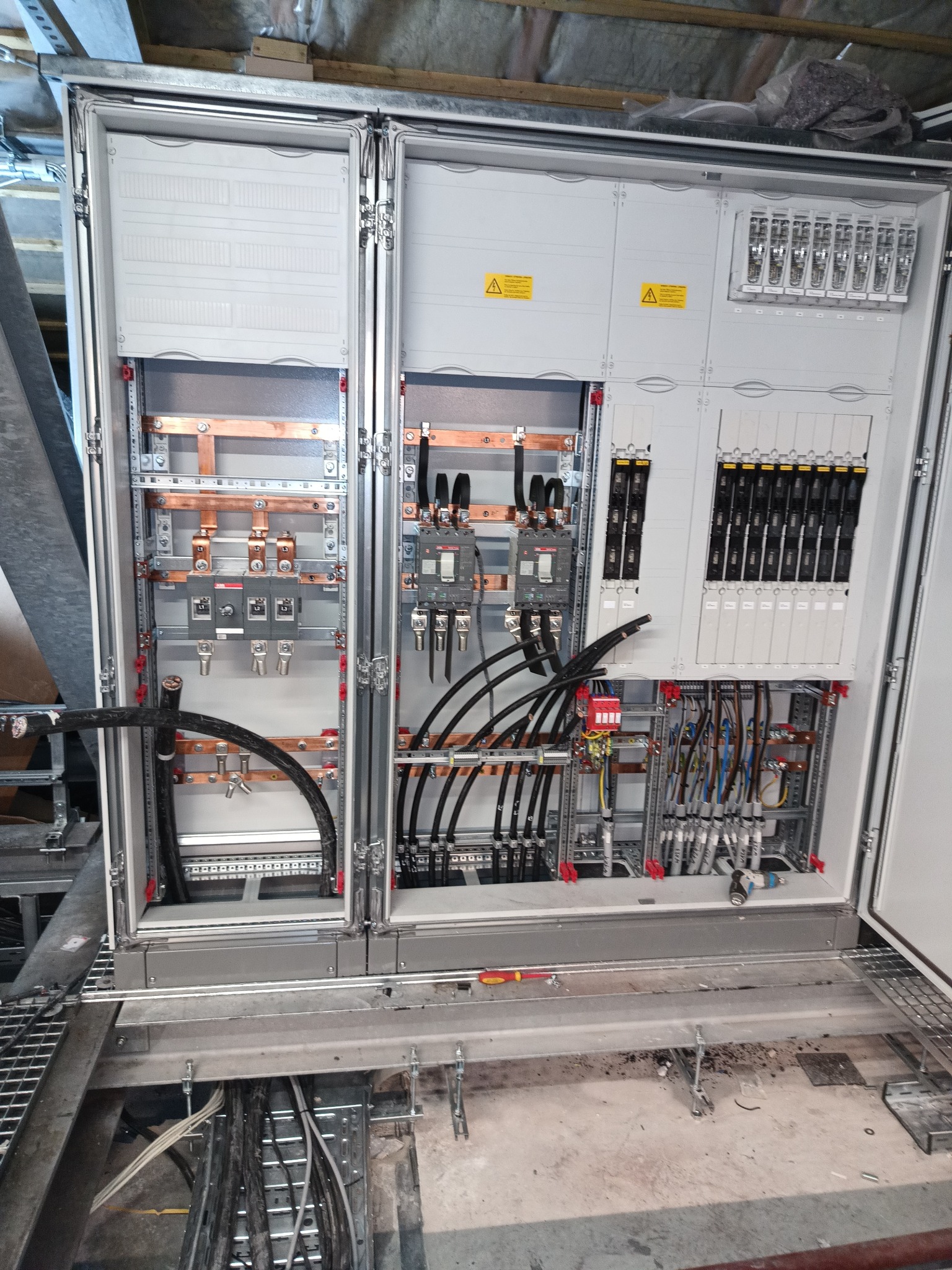 Otwarta szafa elektryczna w trakcie montażu, widoczne kable, szyny, bezpieczniki, styczniki i elementy montażowe, z żółtymi naklejkami ostrzegawczymi.