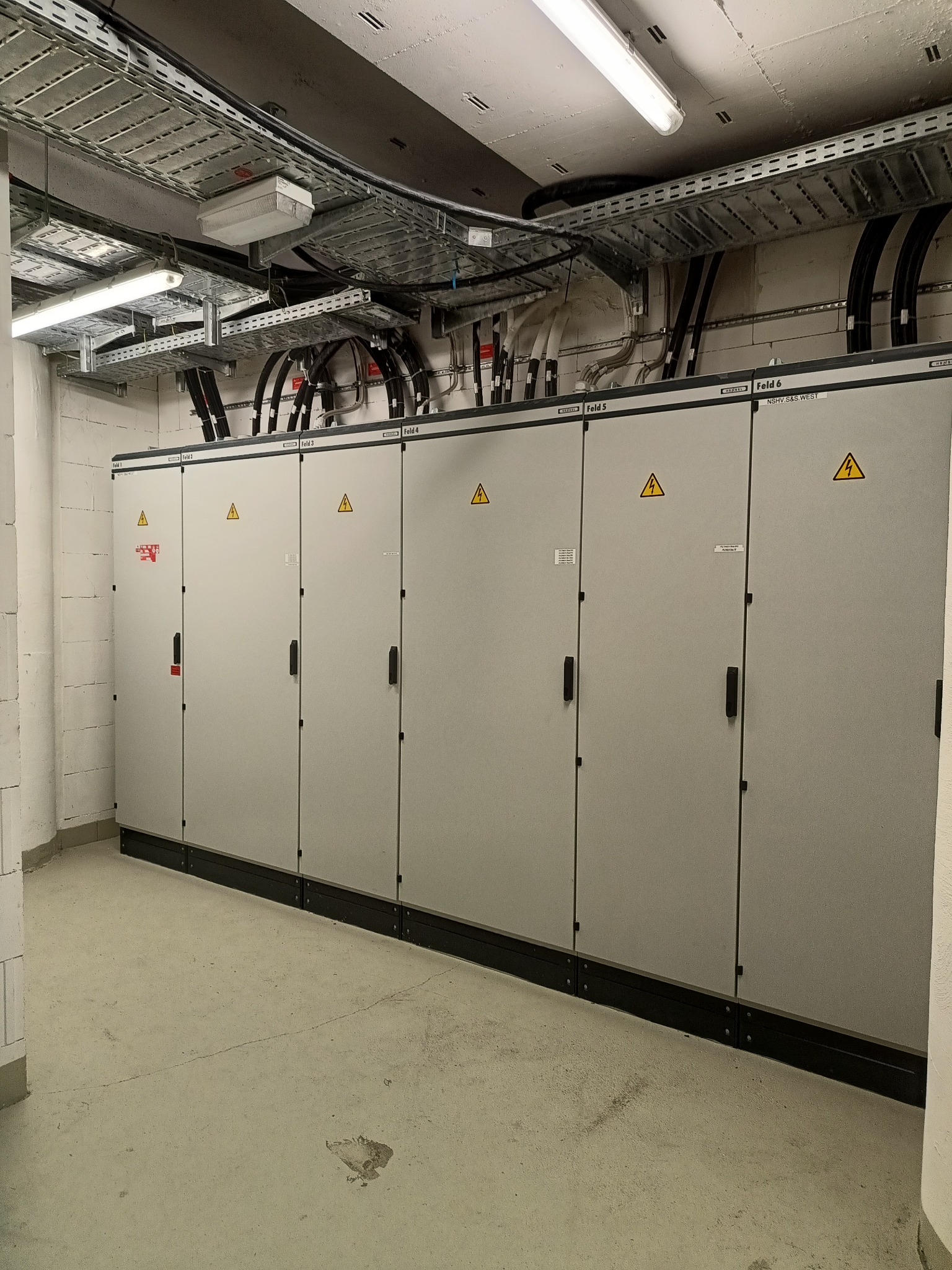 Szereg szaf elektrycznych z ostrzeżeniami o wysokim napięciu, zamontowanych w pomieszczeniu technicznym z widocznymi kablami i kanałami kablowymi pod sufitem.