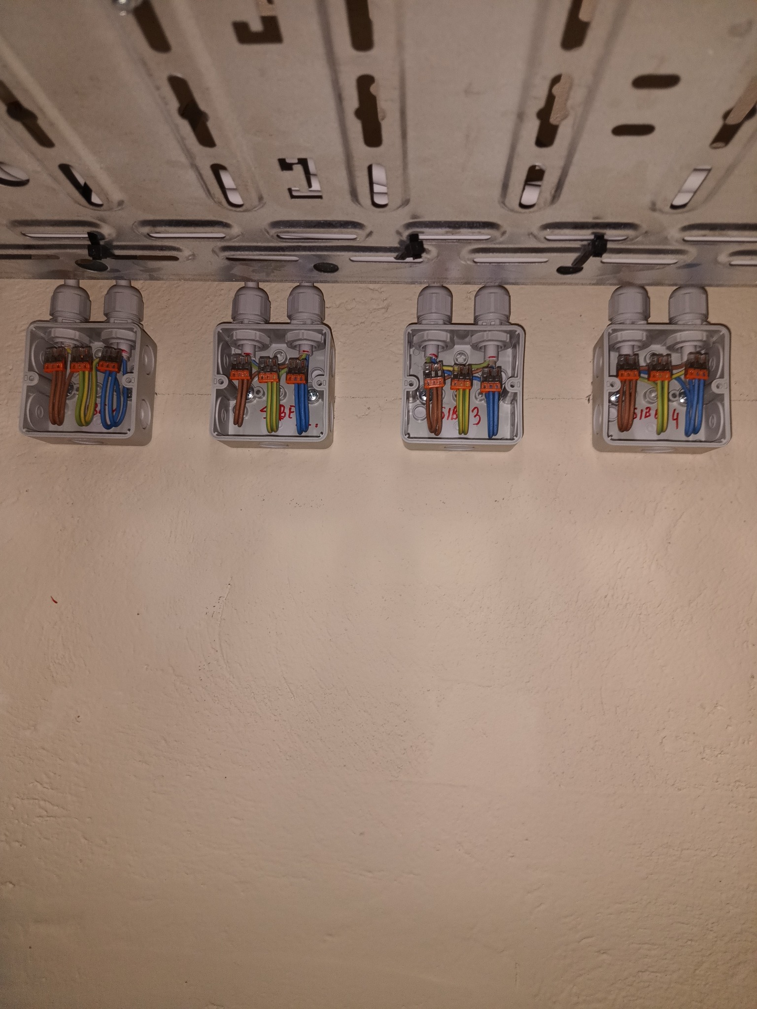 Cztery puszki instalacyjne z podłączonymi przewodami elektrycznymi zamocowane do perforowanej szyny montażowej pod sufitem, widoczne oznaczenia numerów przewodów na puszkach.