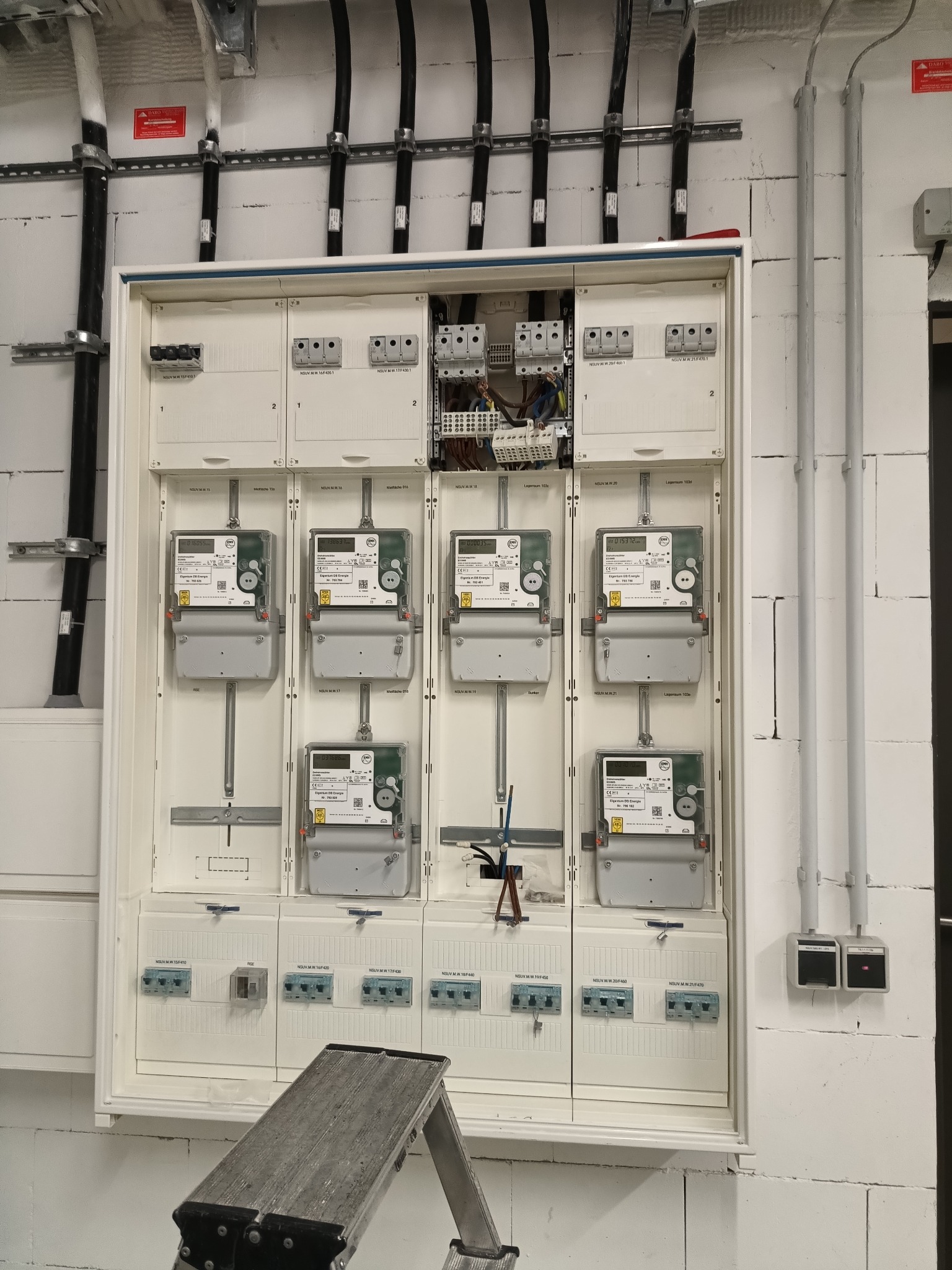 Rozdzielnica elektryczna z zamontowanymi licznikami energii, widoczne kable i przewody, drabina przystawiona do ściany z białej cegły.