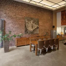 Jasnoszara podłoga w przestronnym wnętrzu z ceglaną ścianą, drewnianym stołem i krzesłami z wikliny, oświetlona dekoracyjnym sufitem z podświetlanych paneli.