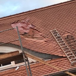 CARBUD - Uszkodzony dach z czerwonej dachówki, częściowo przykryty czerwoną folią ochronną, widoczne rusztowanie i drabina oparta o dach.