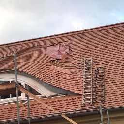 CARBUD - Częściowo uszkodzony dach z czerwonej dachówki karpiówki, zabezpieczony folią w miejscu ubytku, widoczne rusztowanie i drabina dekarska.