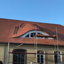 CARBUD - Ceglastoczerwony dach z lukarną w kształcie łuku, fragment dachu z uszkodzeniami, drabina dekarska i rusztowanie przed budynkiem o jasnej elewacji.
