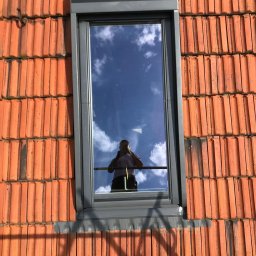 CARBUD - Nowo zamontowane okno dachowe z szarą ramą, widoczne na tle pomarańczowej dachówki; w szybie odbija się niebo i osoba robiąca zdjęcie.