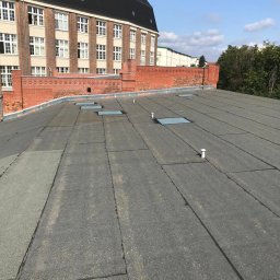 CARBUD - Płaski dach pokryty papą termozgrzewalną z widocznymi świetlikami dachowymi i kominkami wentylacyjnymi na tle ceglanego budynku z oknami.