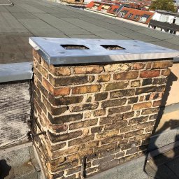 CARBUD - Murowany komin z metalowym daszkiem na płaskim dachu pokrytym papą, widoczne zabudowania w tle.