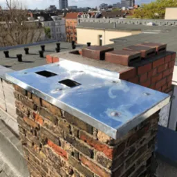 Błyszcząca, metalowa obróbka komina na tle miejskiego krajobrazu, z widocznymi elementami instalacji wentylacyjnej i ceglaną konstrukcją komina.