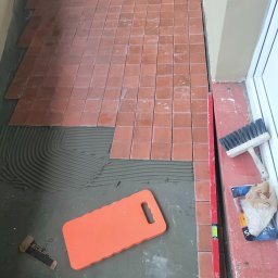 CARBUD - Proces układania terakotowej podłogi z płytek kwadratowych, widoczna warstwa kleju z charakterystycznymi rowkami, pomarańczowa podkładka pod kolana i poziomica przy ścianie.