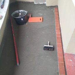 CARBUD - Przygotowanie podłoża z wylewką samopoziomującą, widoczne narzędzia: poziomica, wiadro, pędzel do fugowania i kostka brukowa w rogu pomieszczenia.