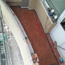 CARBUD - Układanie brązowych płytek ceramicznych na balkonie z widocznymi krzyżykami dystansowymi, poziomica i karton z zapasem płytek.