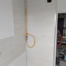 CARBUD - Wykończona ściana w łazience z beżowymi płytkami, widoczna instalacja gazowa z żółtą rurką oraz fragment sylwetki osoby w tle.