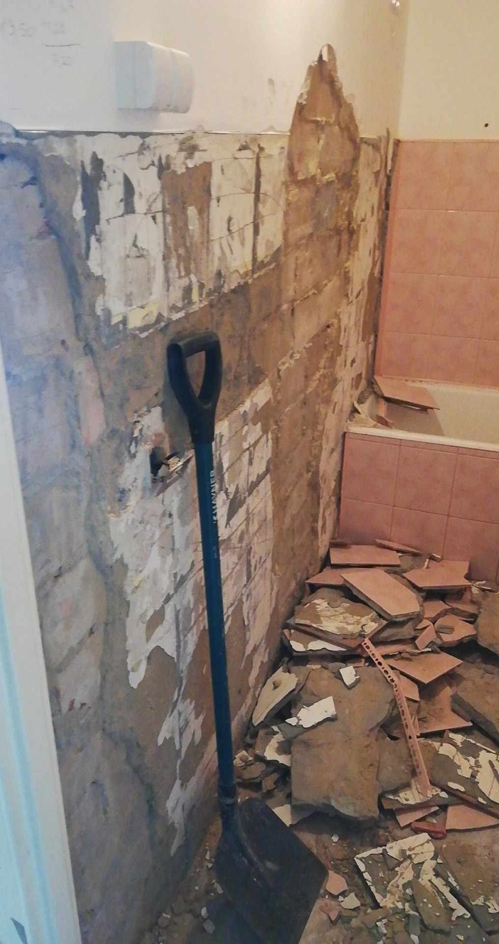 Ściana łazienki w trakcie skuwania starych płytek, widoczna gruzowisko z płytek i łopata oparta o ścianę.