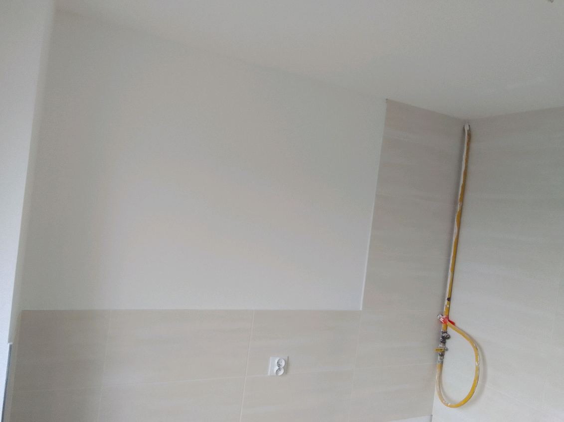 Wykończona ściana w łazience z beżowymi płytkami imitującymi kamień, widoczny fragment instalacji wodnej w żółtej otulinie i gniazdko elektryczne.