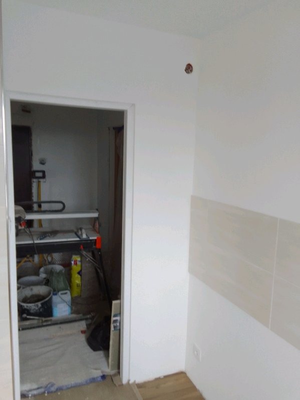 Wnętrze w trakcie remontu: widoczne płytki na ścianie, narzędzia i materiały budowlane w pomieszczeniu obok, zabezpieczona podłoga.