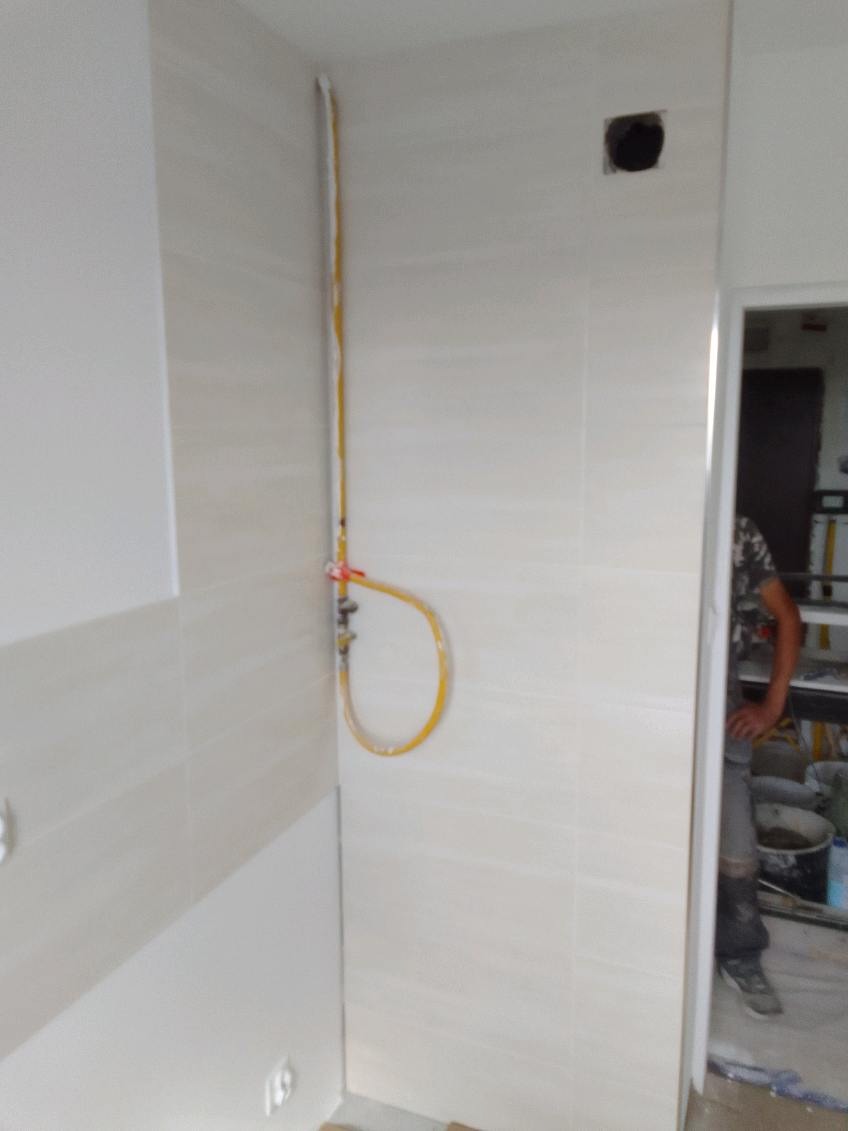 Wykończona ściana w łazience z beżowymi płytkami, widoczna instalacja gazowa z żółtą rurką oraz fragment sylwetki osoby w tle.