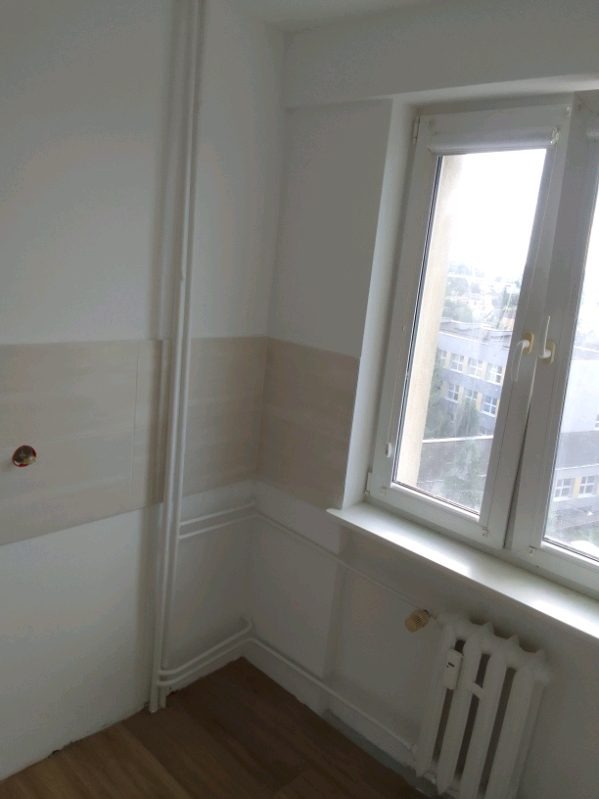 Wykończone wnętrze pokoju z drewnianą podłogą, częściowo wyłożonego jasnymi płytkami, z białym oknem PCV i grzejnikiem żeberkowym