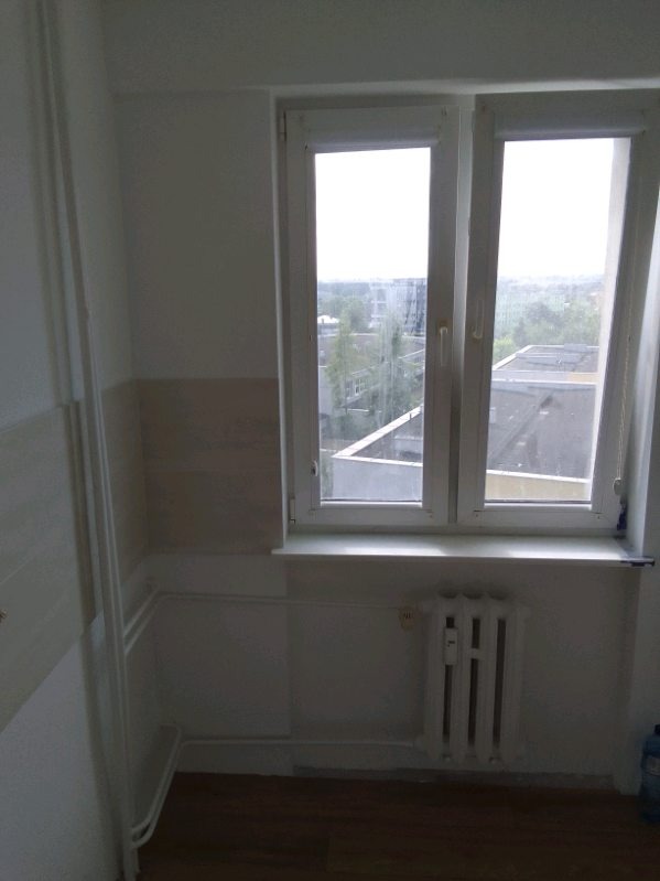 Wnętrze pokoju z oknem PCV, grzejnikiem żeberkowym i panelami ściennymi w odcieniach beżu, widok na zabudowania miejskie za oknem.