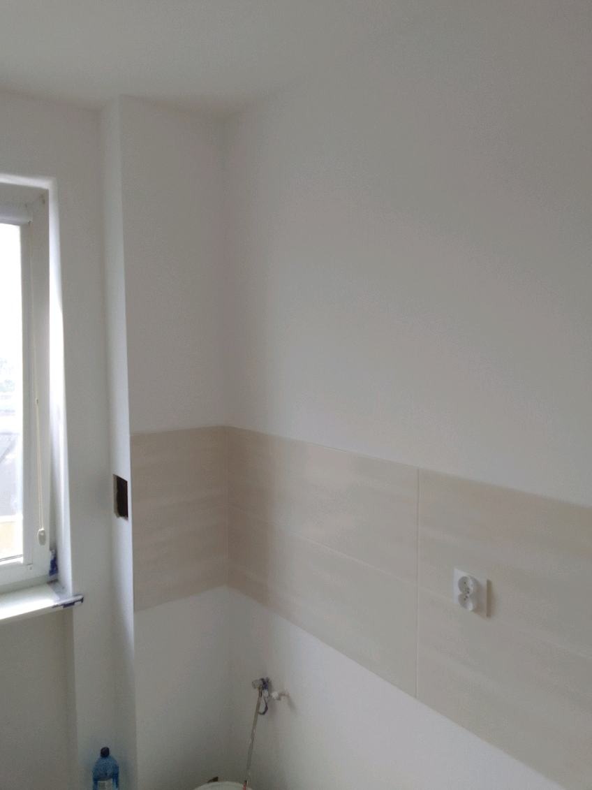 Fragment wykończonej łazienki z beżowymi płytkami ułożonymi do połowy ściany, widoczne gniazdko elektryczne, okno i wystający fragment instalacji wodnej.