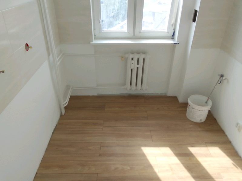 Wnętrze łazienki w trakcie remontu: położona podłoga z paneli imitujących drewno, białe ściany częściowo wykończone płytkami, grzejnik żeberkowy i wiadro z farbą przy ścianie.