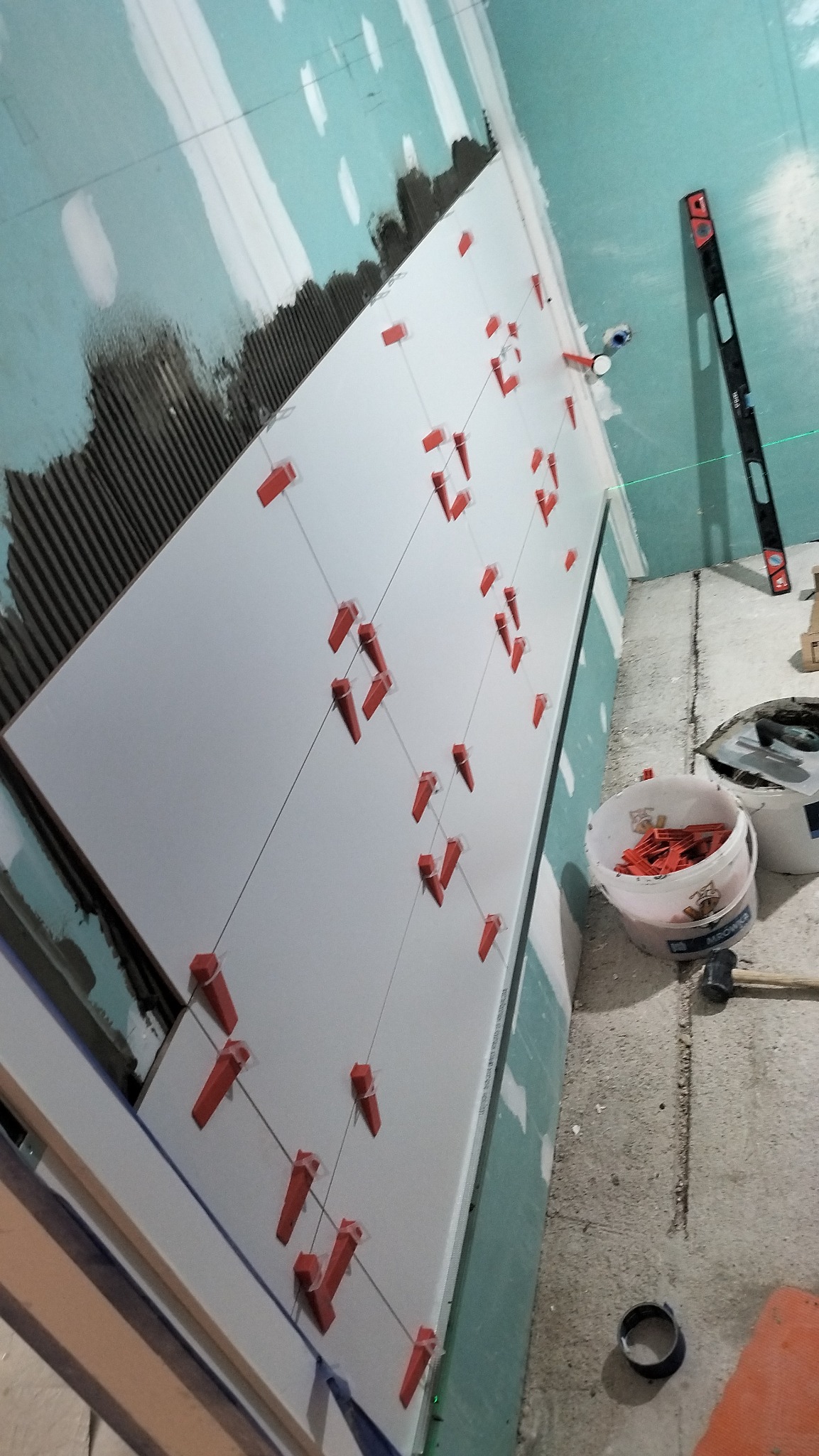 Układanie białych płytek na ścianie z użyciem poziomicy laserowej i czerwonych klinów dystansowych, widoczna struktura kleju i narzędzia pracy.