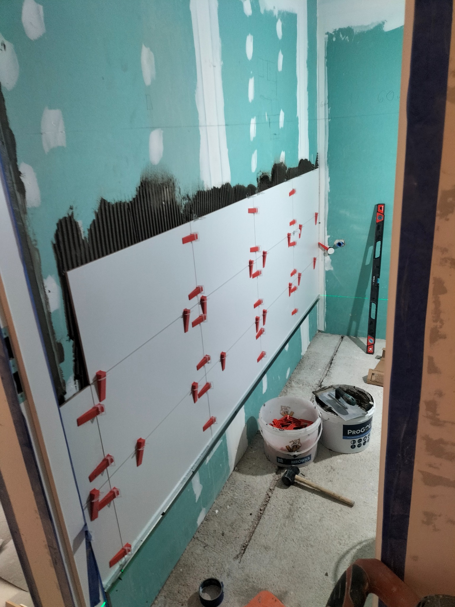 Ściana w trakcie układania białych płytek, widoczne czerwone klipsy poziomujące, klej na ścianie, poziomica laserowa i narzędzia.